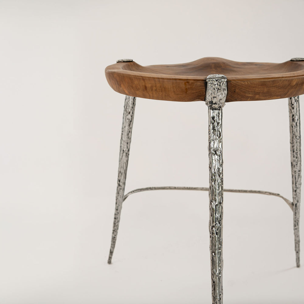 Nahaufnahme eines Barhockers mit einer Sitzfläche aus Teakholz, die natürlich und ergonomisch geformt ist. Die Beine und Verbindungen des Hockers sind aus silbernem Metall mit einer grob texturierten Oberfläche, die eine stilvolle und moderne Optik verleiht.
