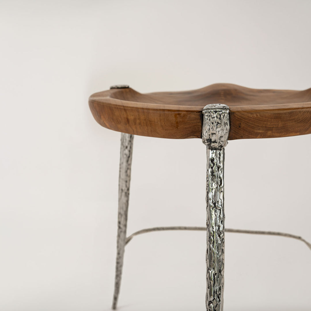 Nahaufnahme eines Barhockers mit einer Sitzfläche aus Teakholz, die natürlich und ergonomisch geformt ist. Die Beine und Verbindungen des Hockers sind aus silbernem Metall mit einer grob texturierten Oberfläche, die eine stilvolle und moderne Optik verleiht.