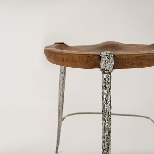 Nahaufnahme eines Barhockers mit einer Sitzfläche aus Teakholz, die natürlich und ergonomisch geformt ist. Die Beine und Verbindungen des Hockers sind aus silbernem Metall mit einer grob texturierten Oberfläche, die eine stilvolle und moderne Optik verleiht.