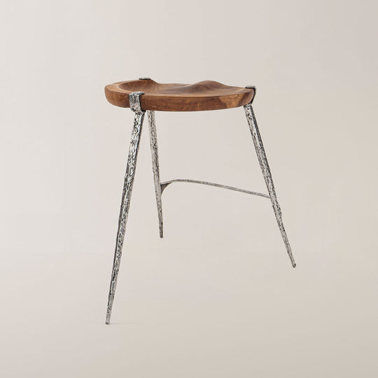 Ein moderner Barhocker mit einer organisch geformten Sitzfläche aus Teakholz und drei eleganten, schlanken Beinen aus poliertem Metall, die ein auffälliges, texturiertes Muster aufweisen. Der Hocker steht vor einem neutralen, hellen Hintergrund, der seine schlichte Eleganz unterstreicht.